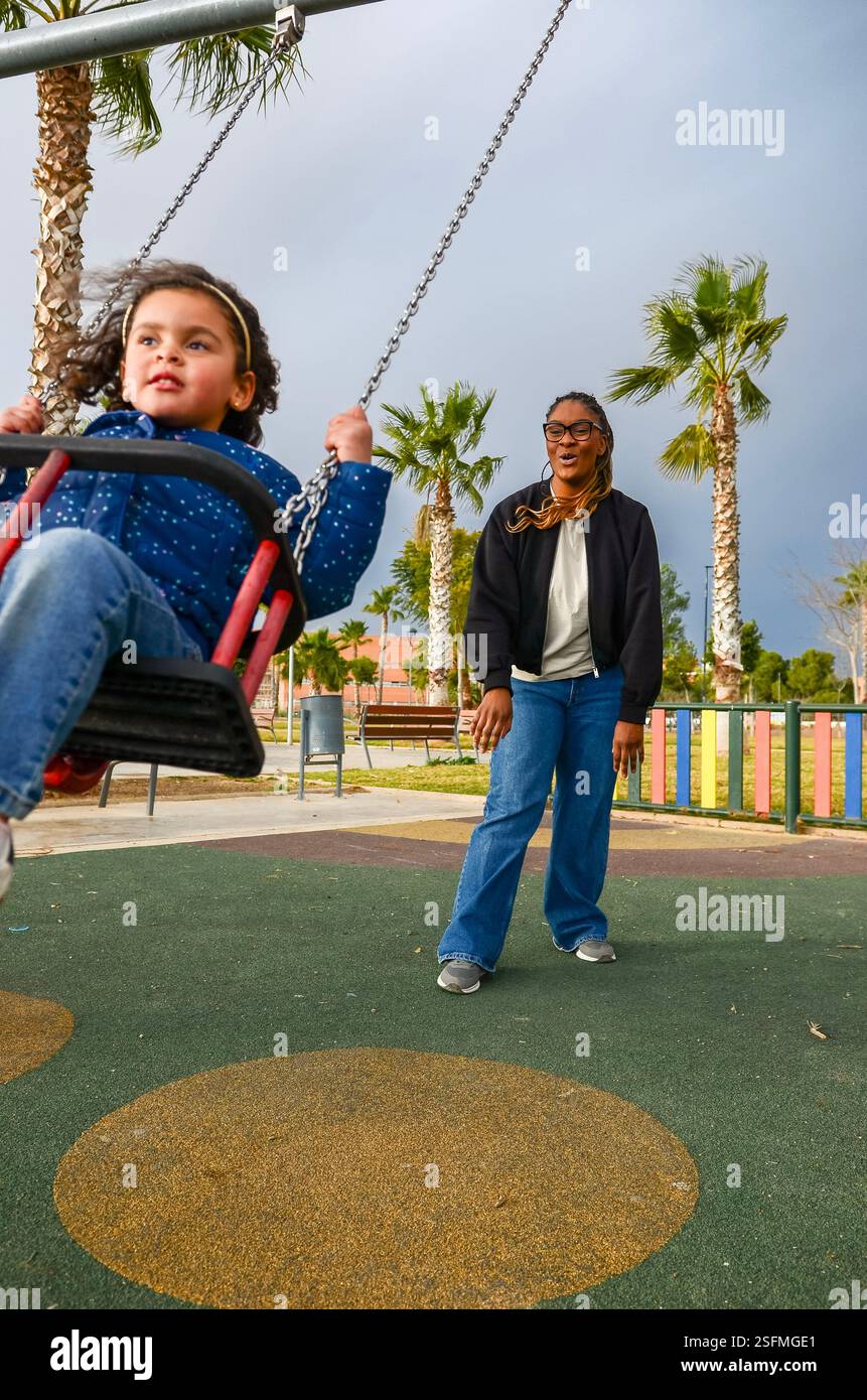 Little girl having fun on a swing pushed by her mother in a playground ...