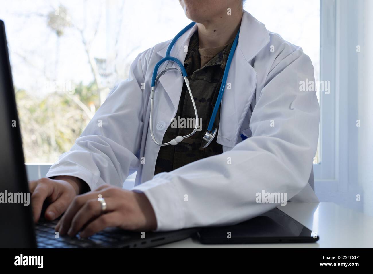 Military doctor in white coat and stethoscope, typing on a laptop ...