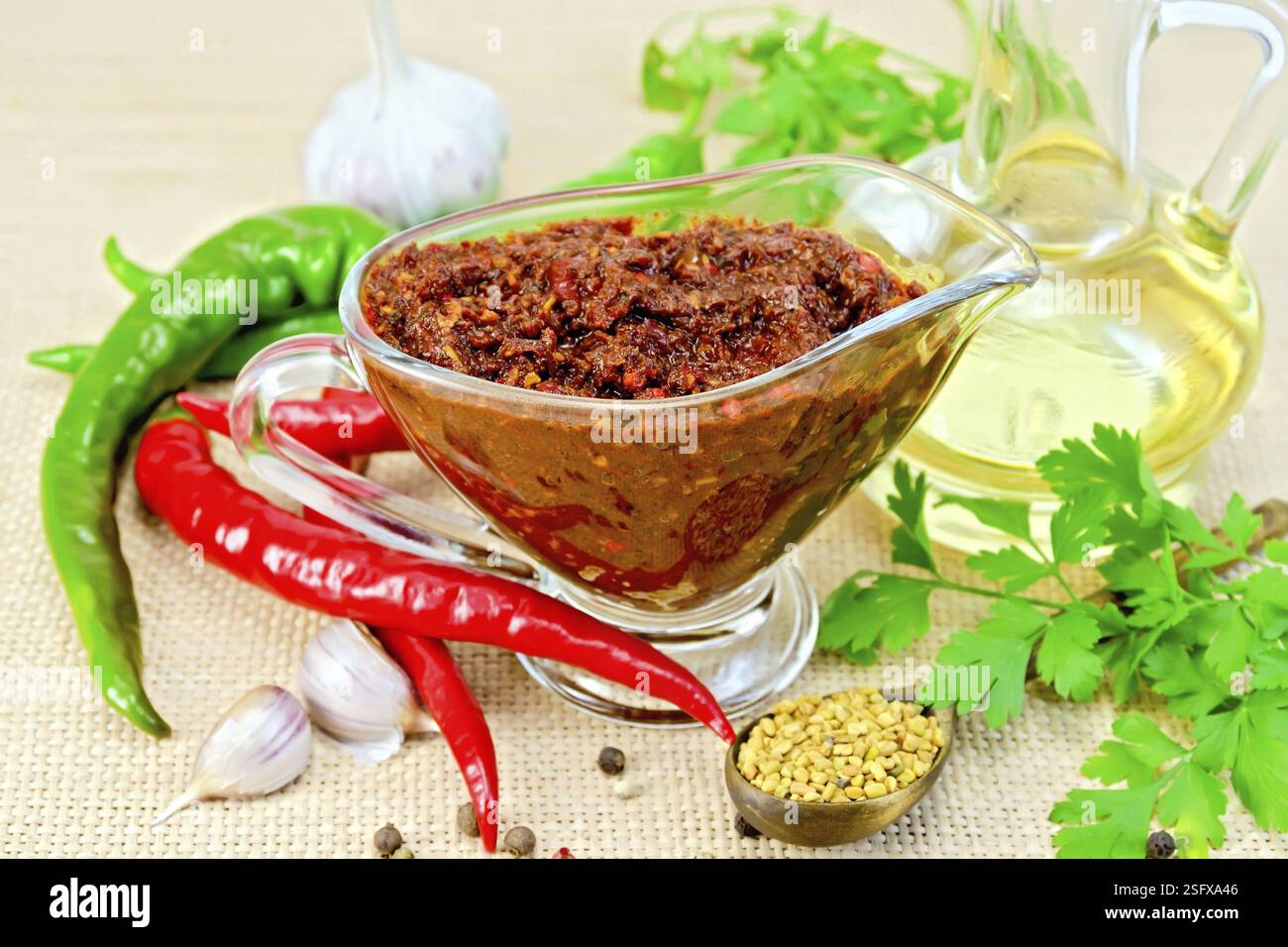 Tabasco adjika in a glass gravy boat, hot pepper, garlic, parsley, fenugreek, oil in the decanter on the background of sack cloth Stock Photo