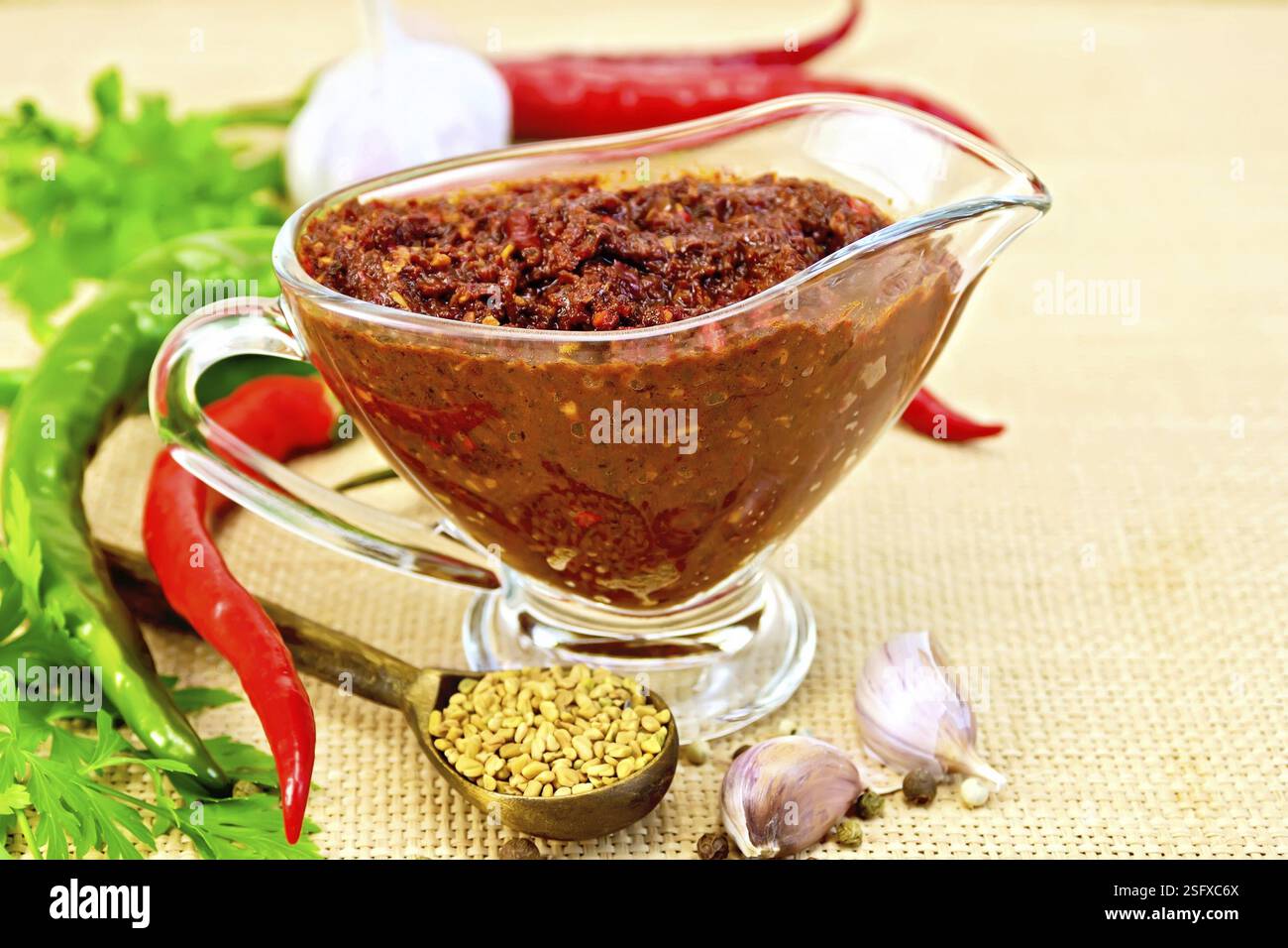 Tabasco adjika in a glass gravy boat, hot pepper, garlic, parsley, fenugreek on the background of sack cloth Stock Photo