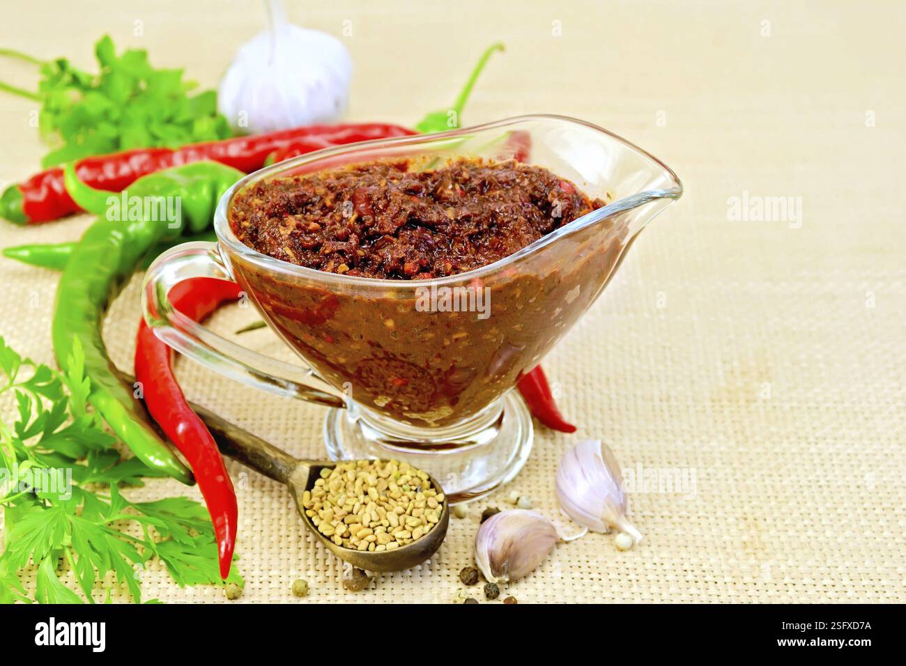 Tabasco adjika in a glass gravy boat, hot pepper, garlic, parsley, fenugreek on the background of coarse cloth sack Stock Photo