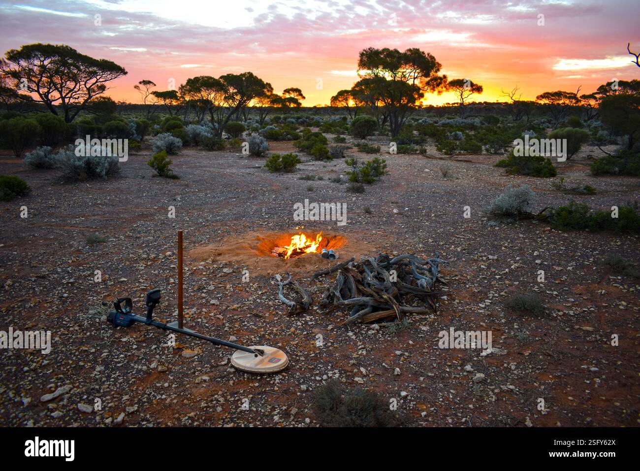 A stunning sunset over the Western Australian desert, featuring a campfire, a pickaxe, and a metal detector in the foreground. Stock Photo