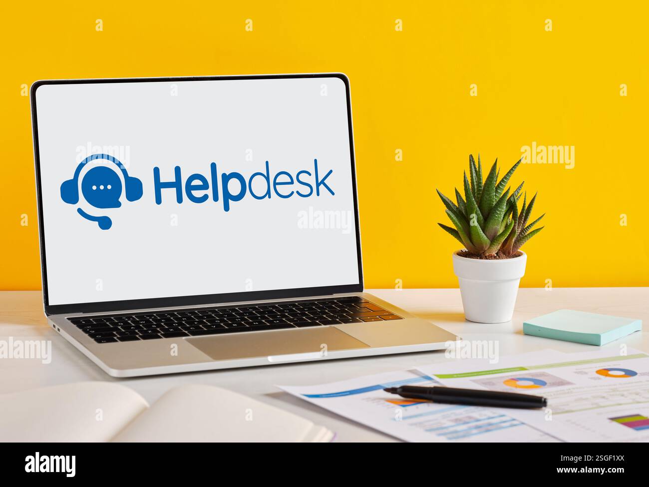 Modern laptop displaying helpdesk software interface on a desk with documents, charts, pen and plant, on a yellow background. Stock Photo