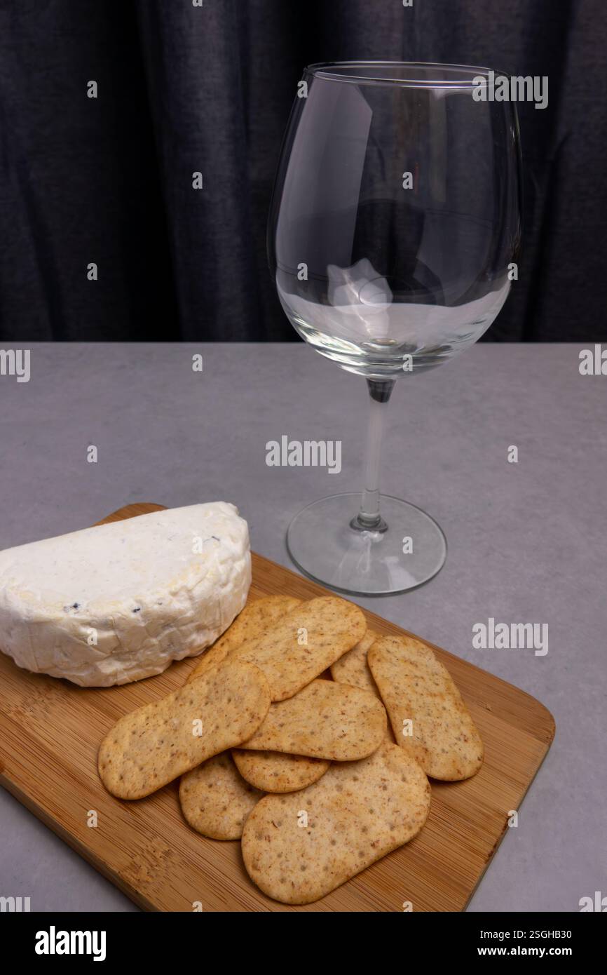 A wooden platter holds soft cheese and crispy crackers next to a clear glass. The backdrop is dark and elegant. Stock Photo