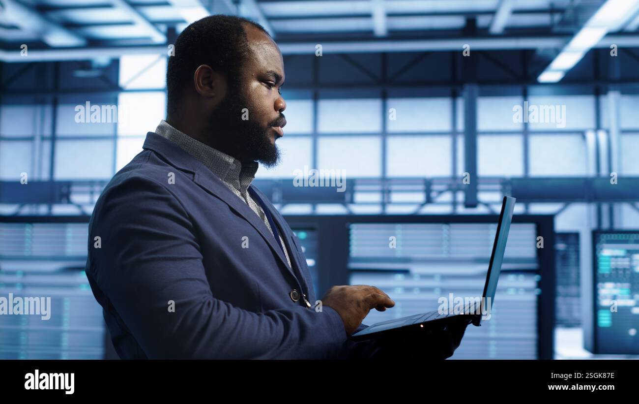 African american man walking in high tech facility, using laptop to patch network issues leading to slow data transfer rates. Data center worker fixing errors affecting supercomputers, camera A Stock Photo