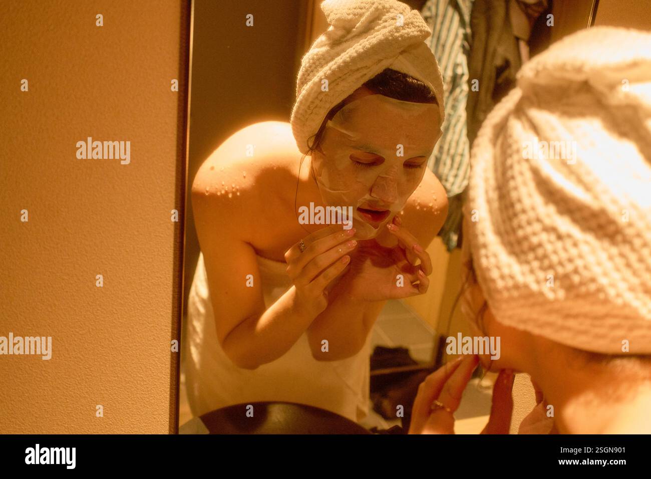 Woman in towel applies a face mask while looking in the mirror in a softly lit hotel bathroom. Tokyo, Japan Stock Photo