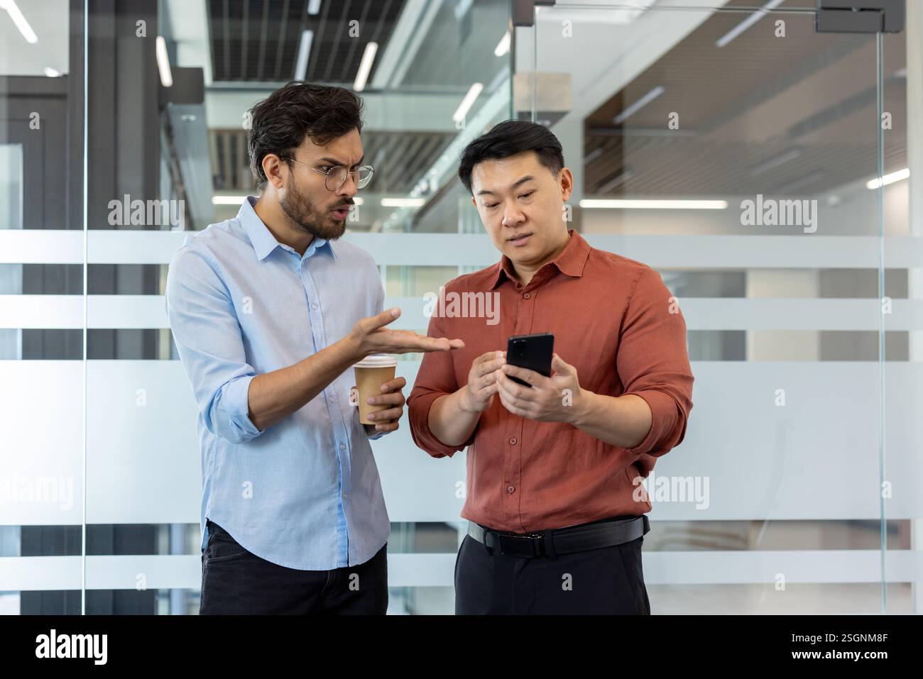 Two colleagues debating and analyzing data on a smartphone while standing in a professional workspace Stock Photo