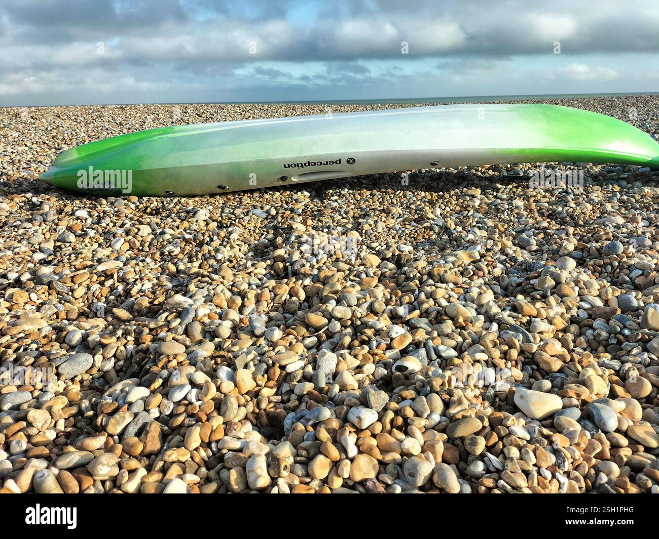 Kayak resting on a pebble beach by the coastline during a cloudy day. A green and white kayak is positioned on a pebble-strewn beach. - Smartphone Captured Stock Image