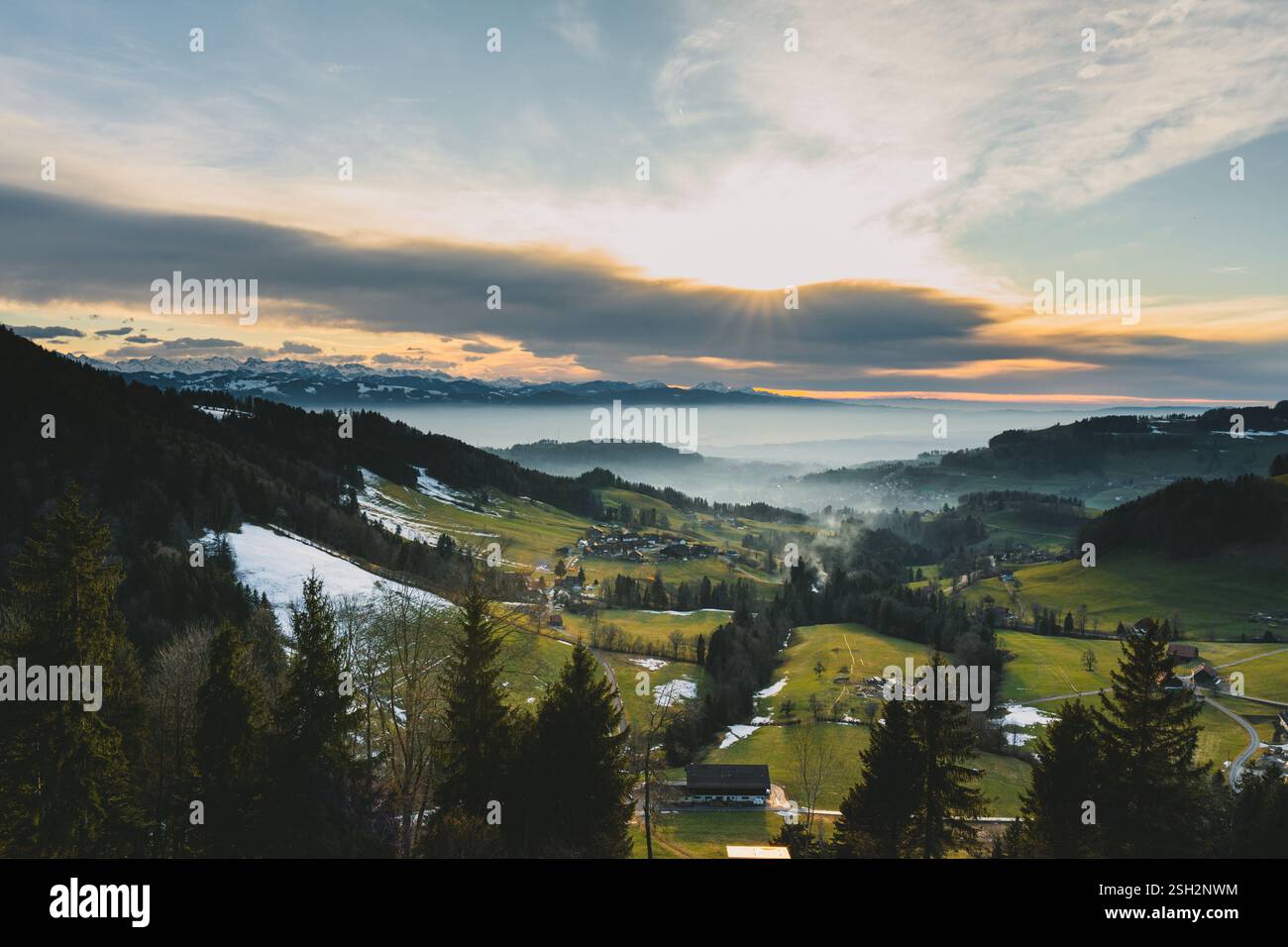 the sun sets behind the majestic mountain range, golden rays pierce through the layered clouds, casting a warm glow over the valley below Stock Photo