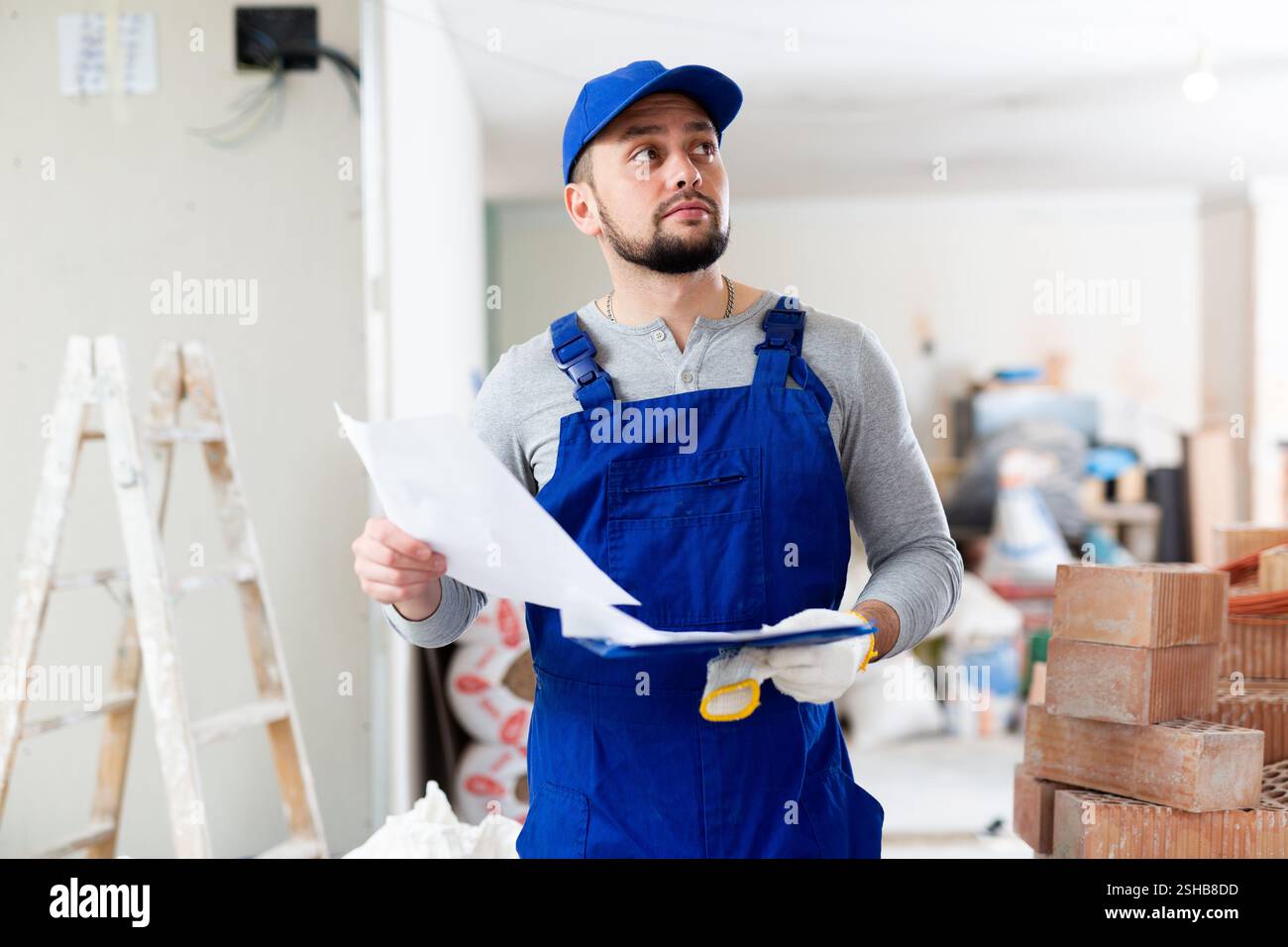Responsible focused bearded supervisor wearing blue overall taking notes on papers while inspecting construction site Stock Photo