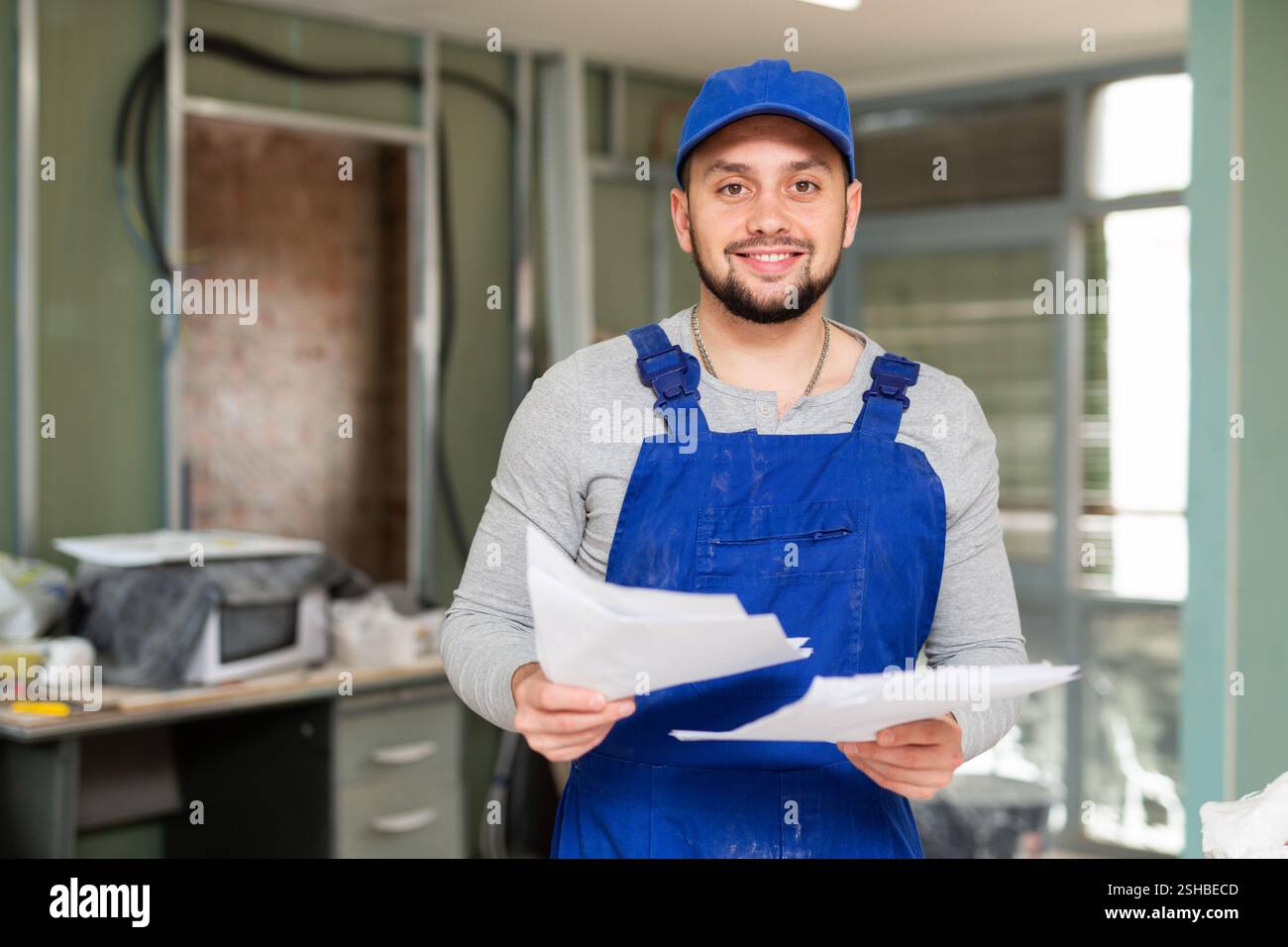 Responsible focused bearded supervisor wearing blue overall taking notes on papers while inspecting construction site Stock Photo