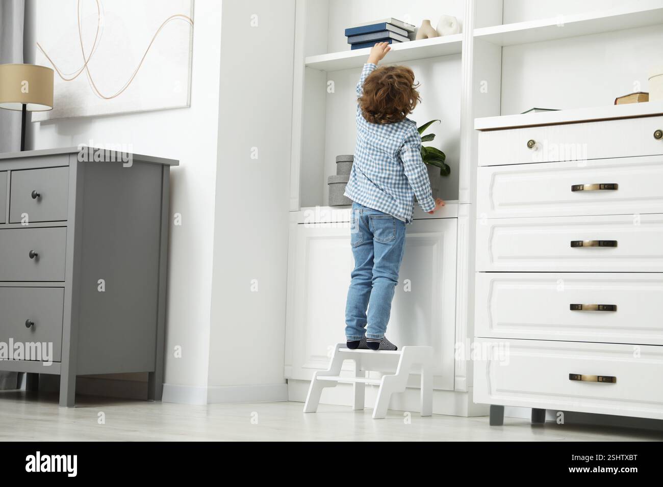Little boy standing on step stool and reaching for book at home, back ...