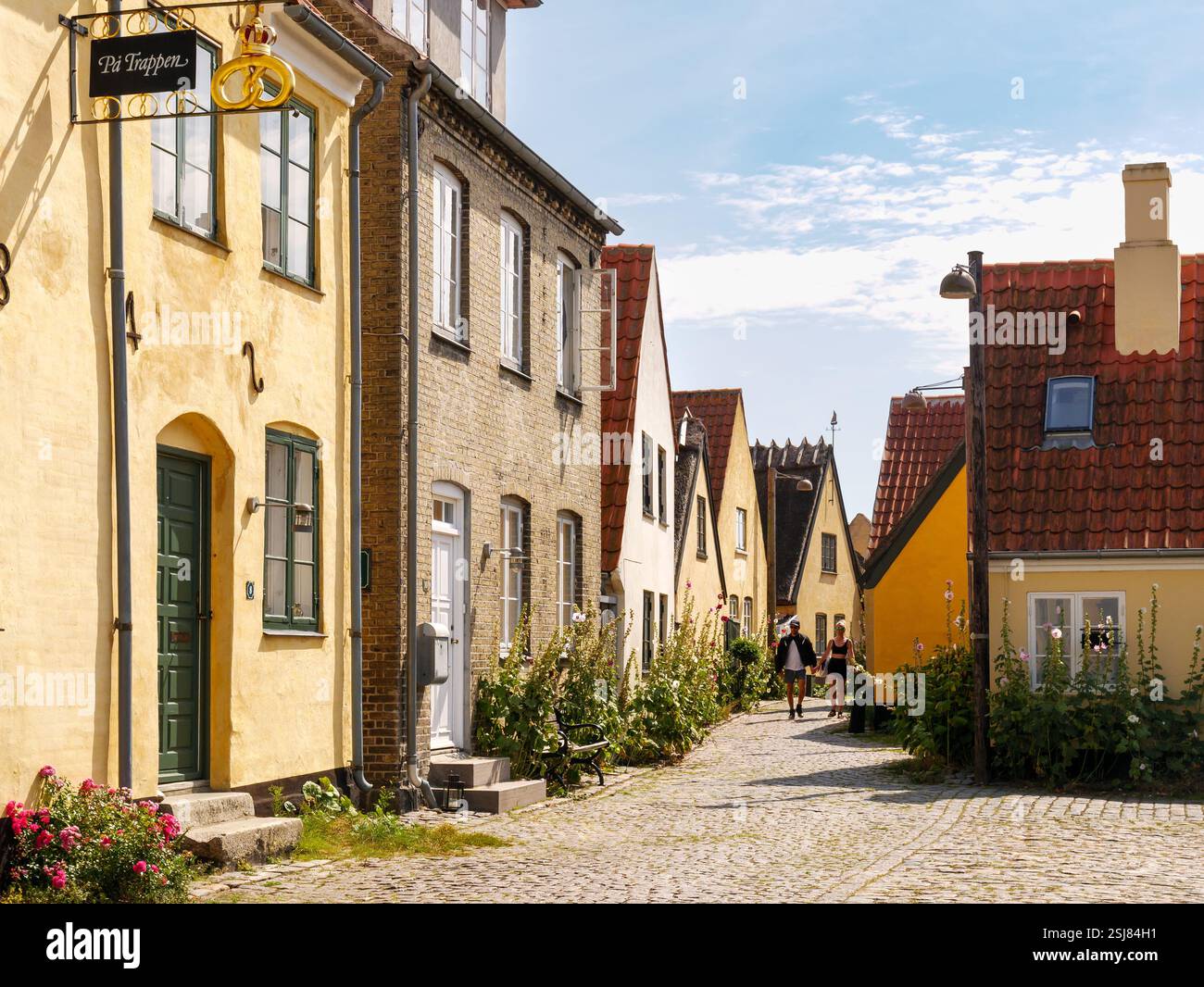 People walking in narrow street lined with hollyhocks and historic ...