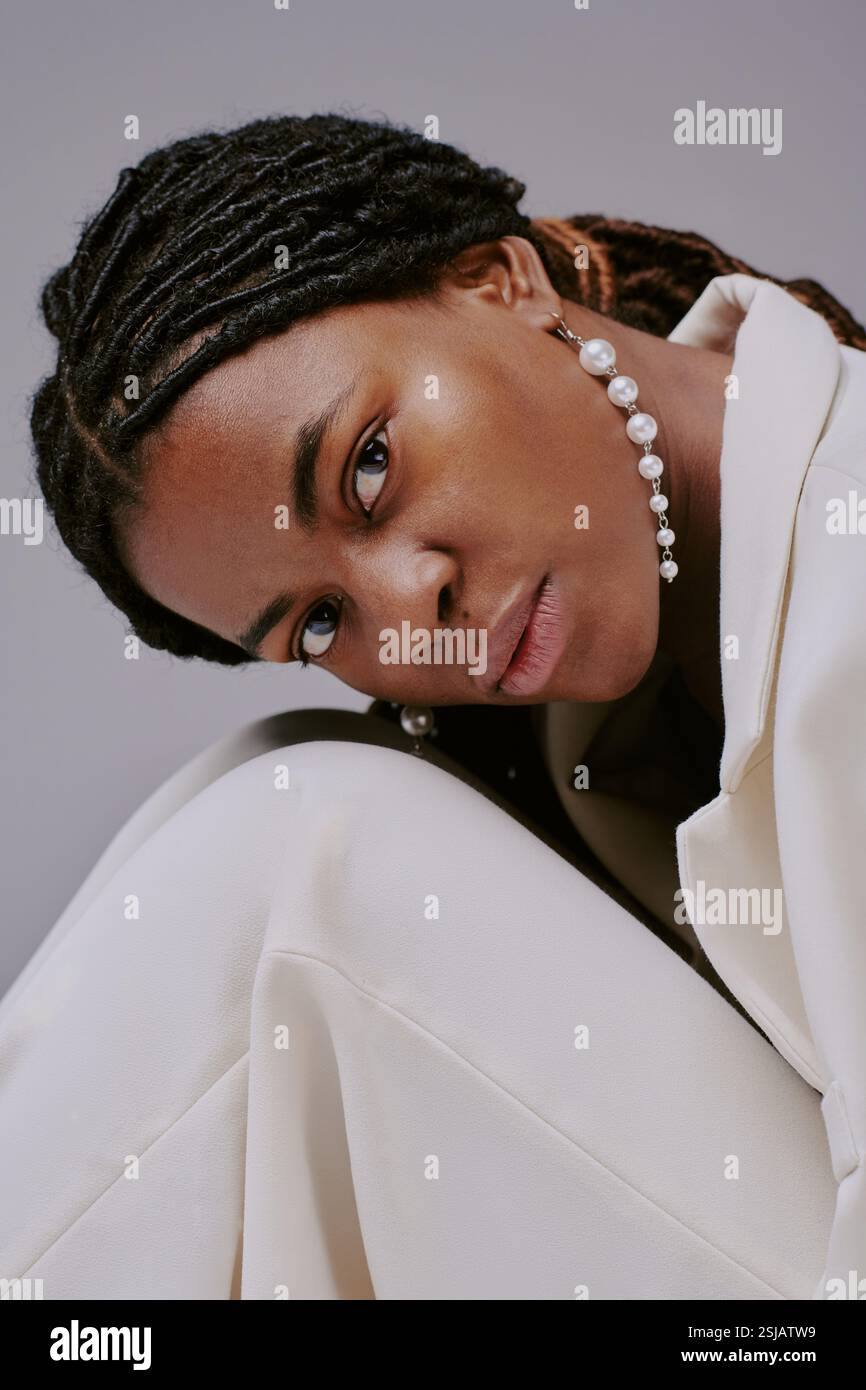 Portrait of woman with braided hair leaning sideways, wearing elegant pearl necklace and light-colored outfit, looking directly at camera Stock Photo