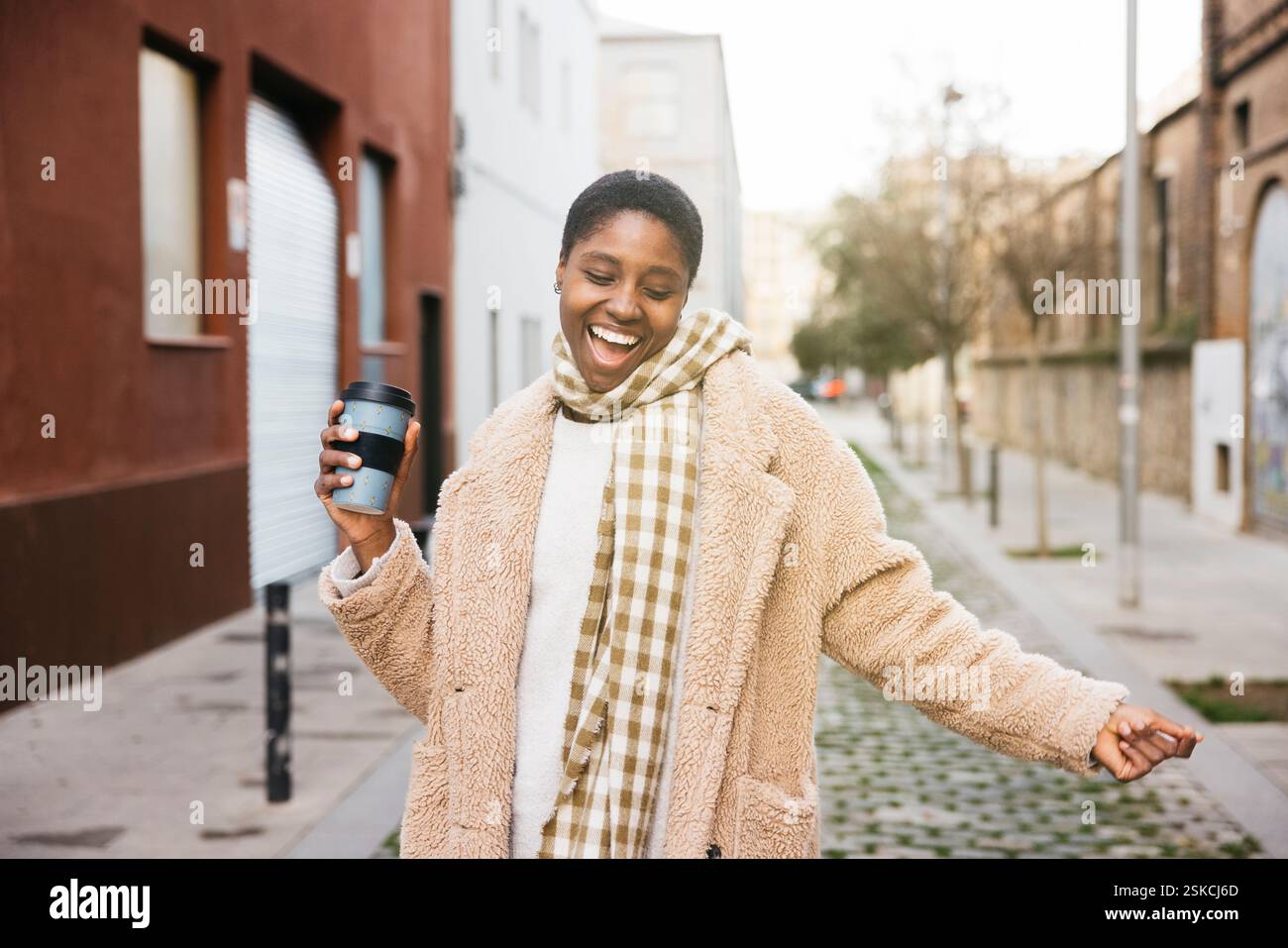 Carefree young woman laughing and holding a reusable coffee cup while walking in the street Stock Photo