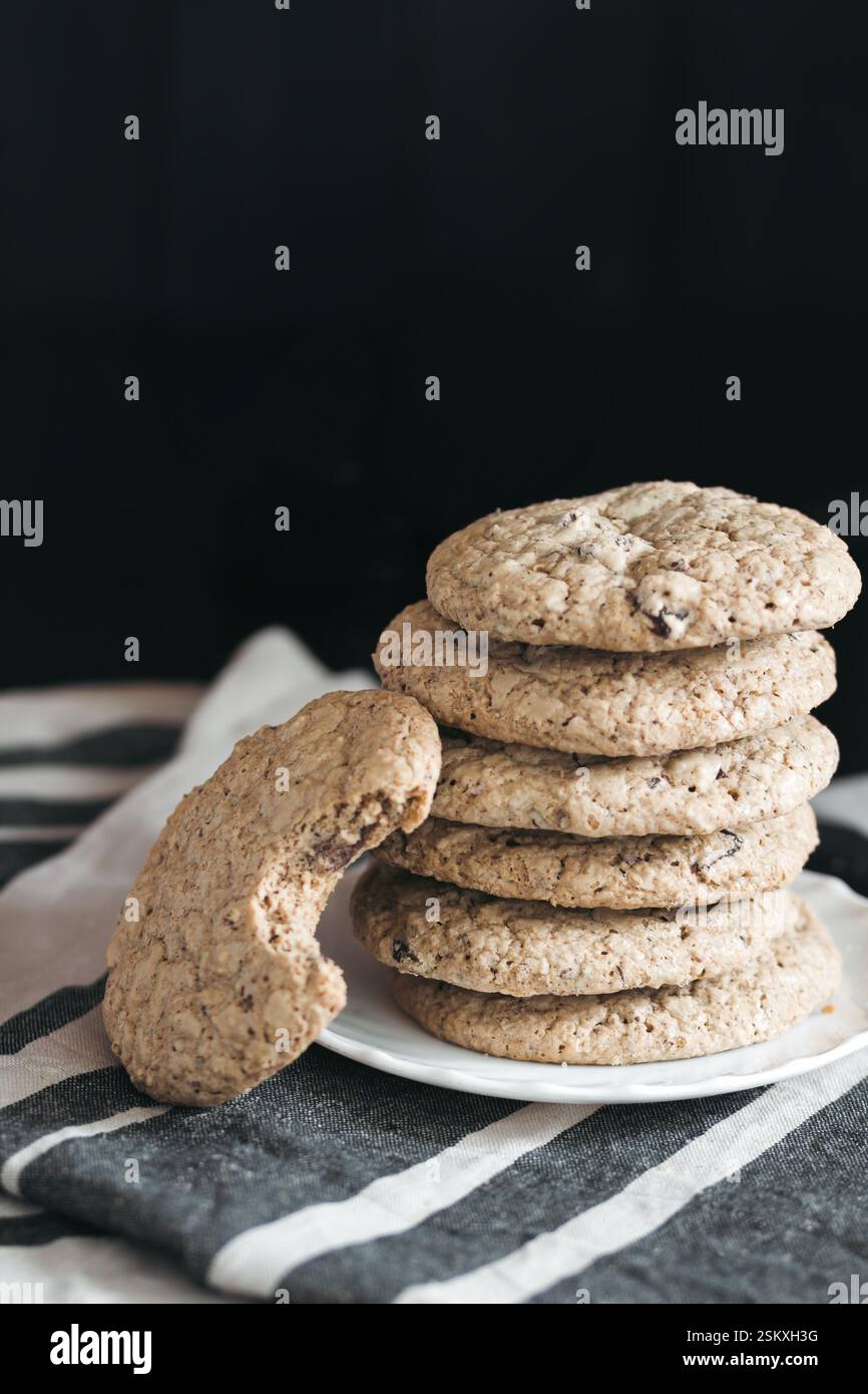 A plate of gluten-free chocolate chip cookies stacked neatly, with one cookie showing a bite taken out, placed on a striped cloth against a dark backg Stock Photo