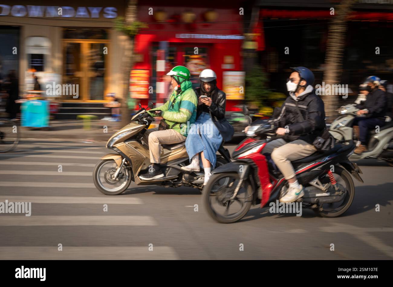 A young Vietnamese woman looks at her mobile smartphone as she rides side saddle on the back of a Grab motorcycle taxi next to Hoan Kiem Lake in centr Stock Photo