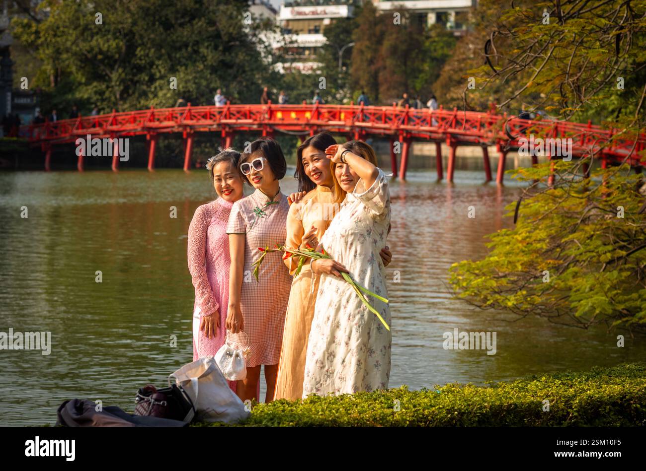 Four Vietnamese women are dressed up and pose for photos against a backdrop of The Huc bridge which leads to Ngoc Son temple on Hoan Kiem Lake, Hanoi, Stock Photo