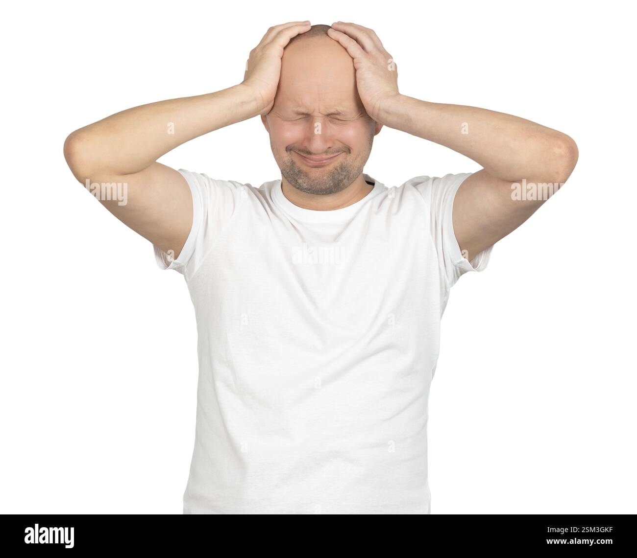 A bald Caucasian man in a white t-shirt stands against a neutral background. He presses both hands against his head, squeezing his eyes shut in pain. Stock Photo
