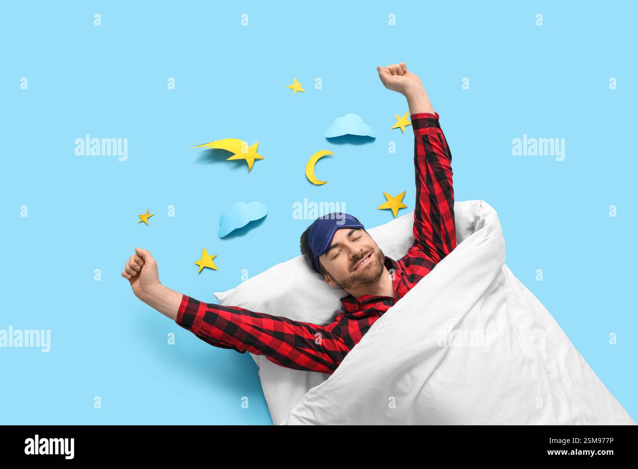 Handsome young man with sleep mask stretching in bed on blue background Stock Photo