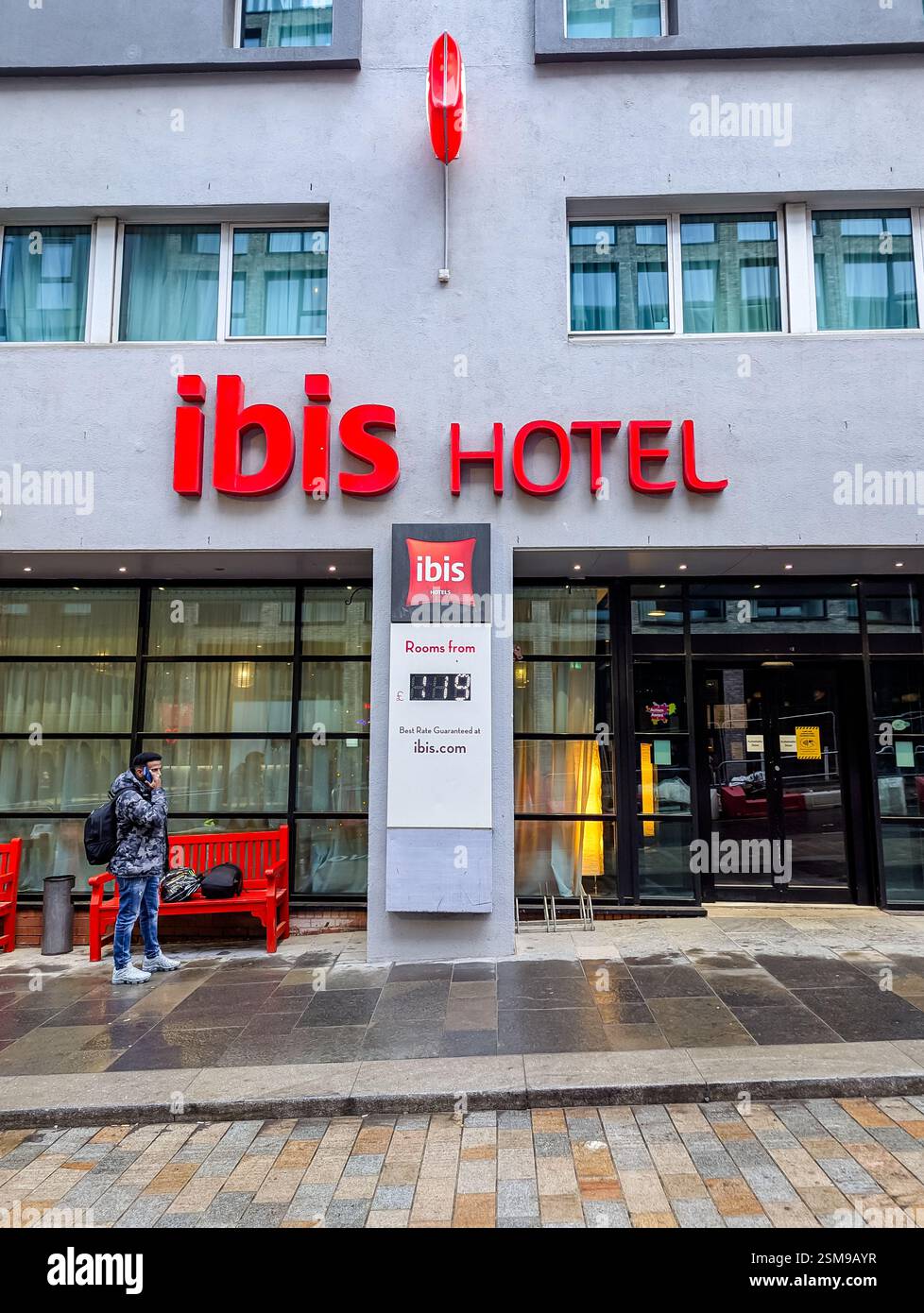 The entrance of an ibis hotel, featuring the hotel's logo in red above the door and a sign indicating rooms are available, with a man standing outside - Smartphone Captured Stock Image