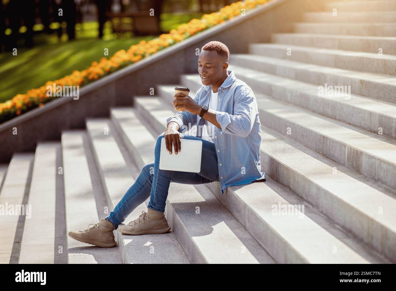 Positive black guy with tablet computer drinking takeaway coffee on stone stairs downtown Stock Photo