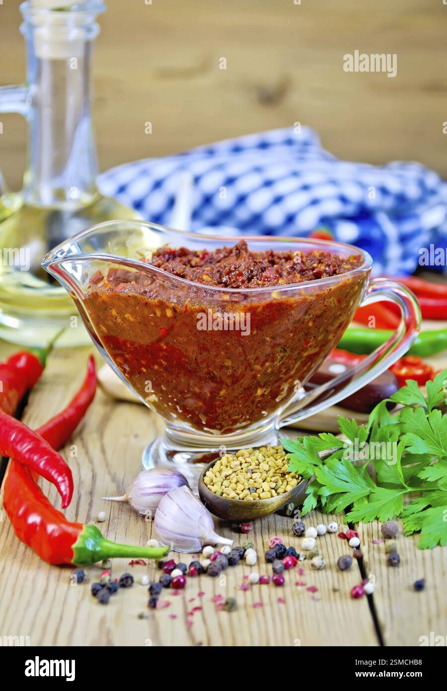 Tabasco adjika in a glass gravy boat, hot pepper, garlic, parsley, fenugreek, napkin, oil in a carafe on a wooden board Stock Photo
