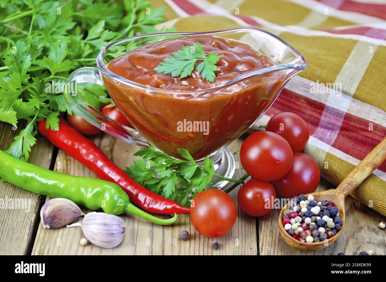 Ketchup in a glass gravy boat, tomatoes, parsley, hot pepper, garlic, napkin on a wooden boards background Stock Photo