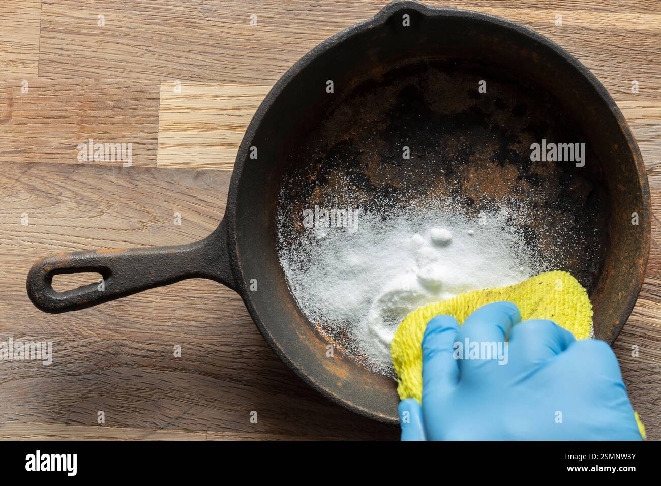 Person cleaning a rusty old cast iron frying pan using bicarbonate of soda and a scouring pad. On a wood board background. Stock Photo