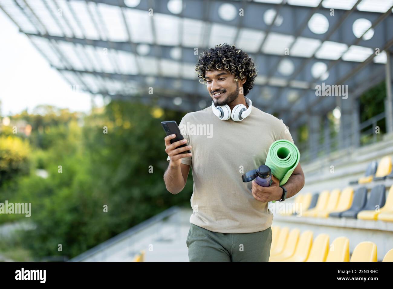 A man with a phone and a yoga mat walks in the park, an athlete uses an application on a smartphone for joint training and acquaintances, books a place in a training class. Stock Photo