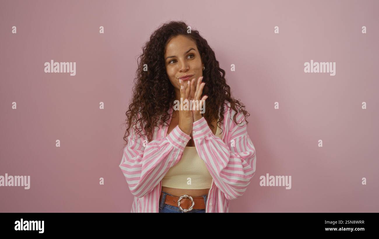 Young woman with curly brunette hair wearing a pink striped shirt and jeans, standing against an isolated pink background, expressing thoughtful and a Stock Photo