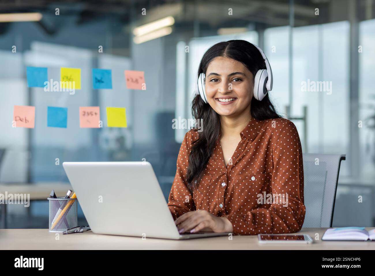 Young woman working inside office wearing headphones, programmer smiling and looking at camera, employee coding new software, developing applications. Stock Photo