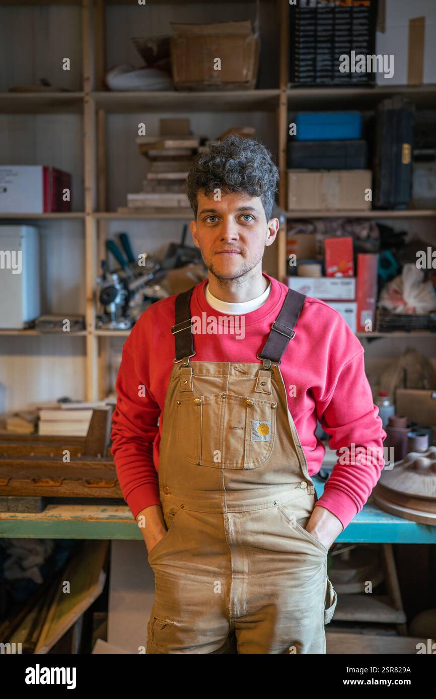 Smiling man carpenter resting in workshop looking at camera putting hands in pockets leans on table Stock Photo