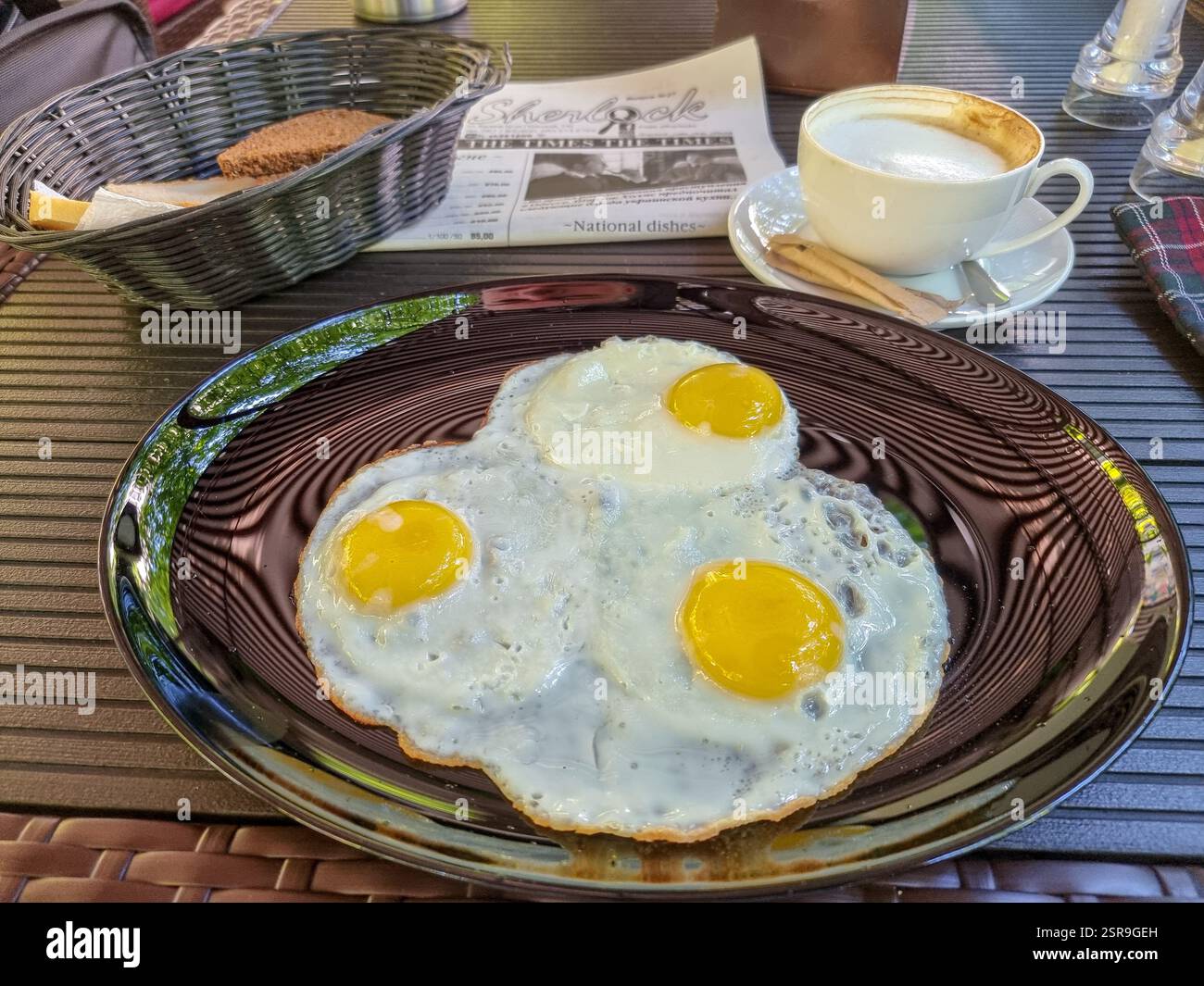 Three fried eggs on a shiny plate, a basket of bread and a cappuccino as breakfast - Smartphone Captured Stock Image