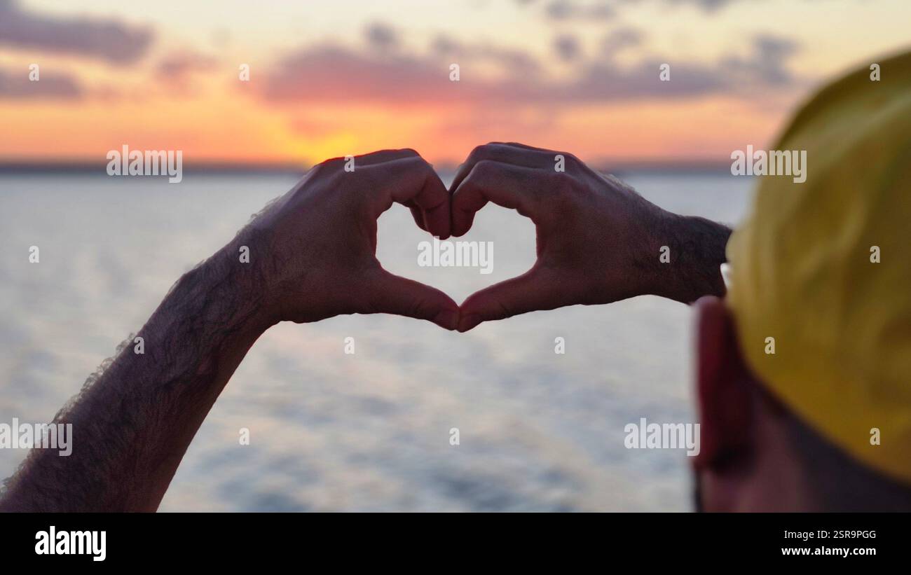 Silhouette of hands forming a heart against a vivid sunset over a vast body of water, evoking emotions of love, warmth, and tranquility as clouds floa - Smartphone Captured Stock Image