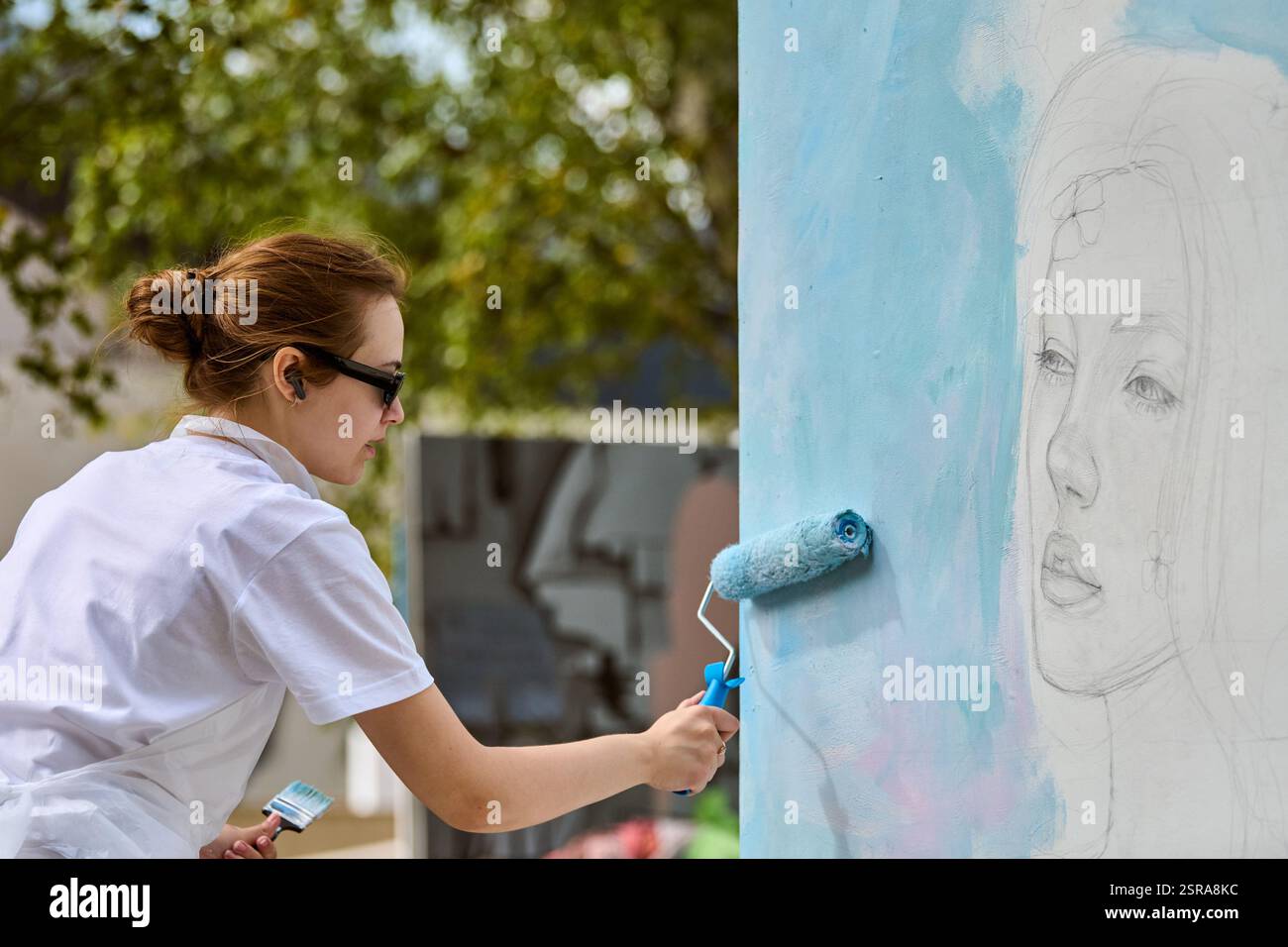 Young female artist painting mural on wall using roller. Outdoor ...