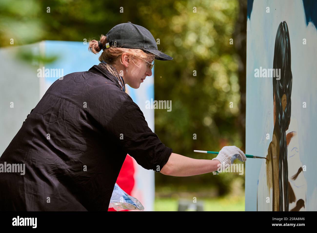 Young female artist with cap and gloves painting on large canvas in ...