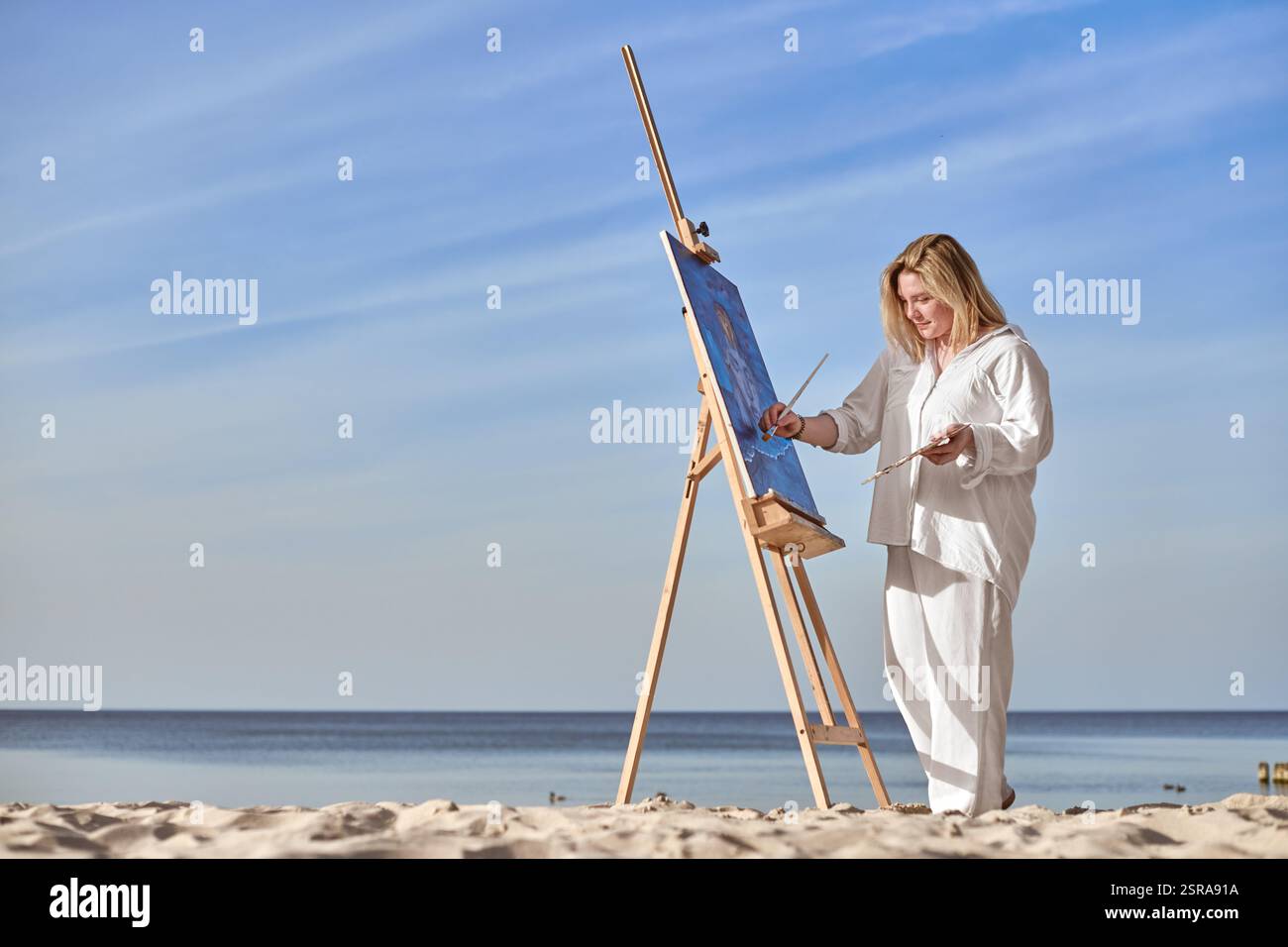 Young woman artist stands near ocean, painting on easel. Serene beach ...
