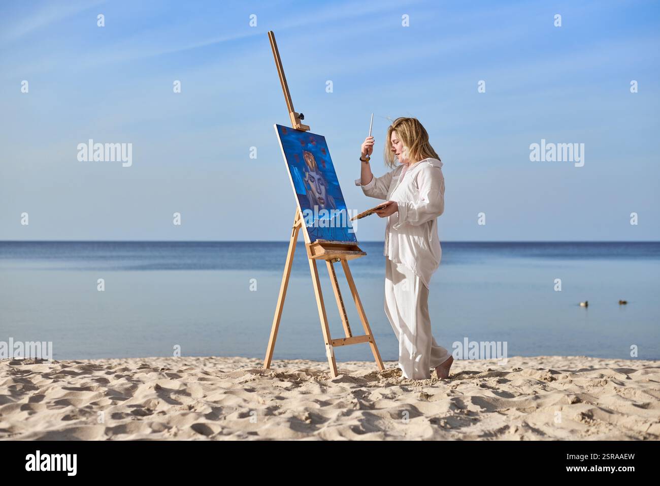 Young woman artist stands near ocean, painting on easel. Serene beach ...