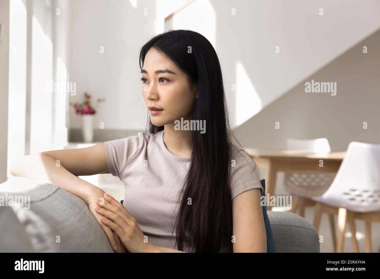 Serious Asian young woman sits thoughtfully on comfortable sofa Stock Photo