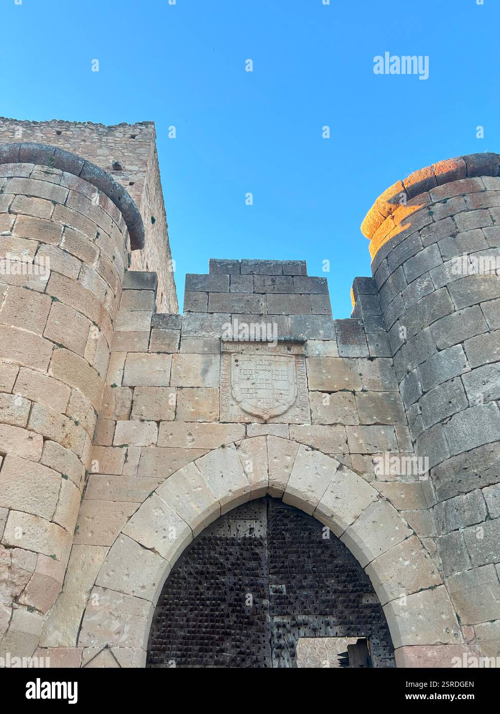 Front of medieval castle facade with stone coat of arms, twin towers, and large wooden iron-spiked gate, reflecting historic European architecture - Smartphone Captured Stock Image