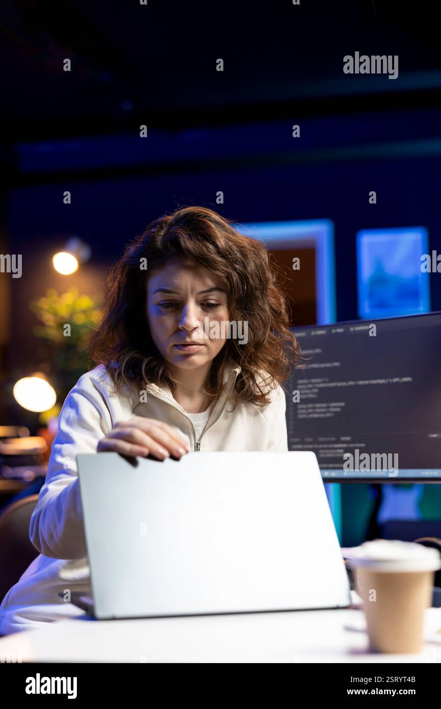 IT expert working from home, opening laptop, testing and deploying programs and systems. Engineer checking code displayed on computer monitors, sitting at desk chair in apartment Stock Photo