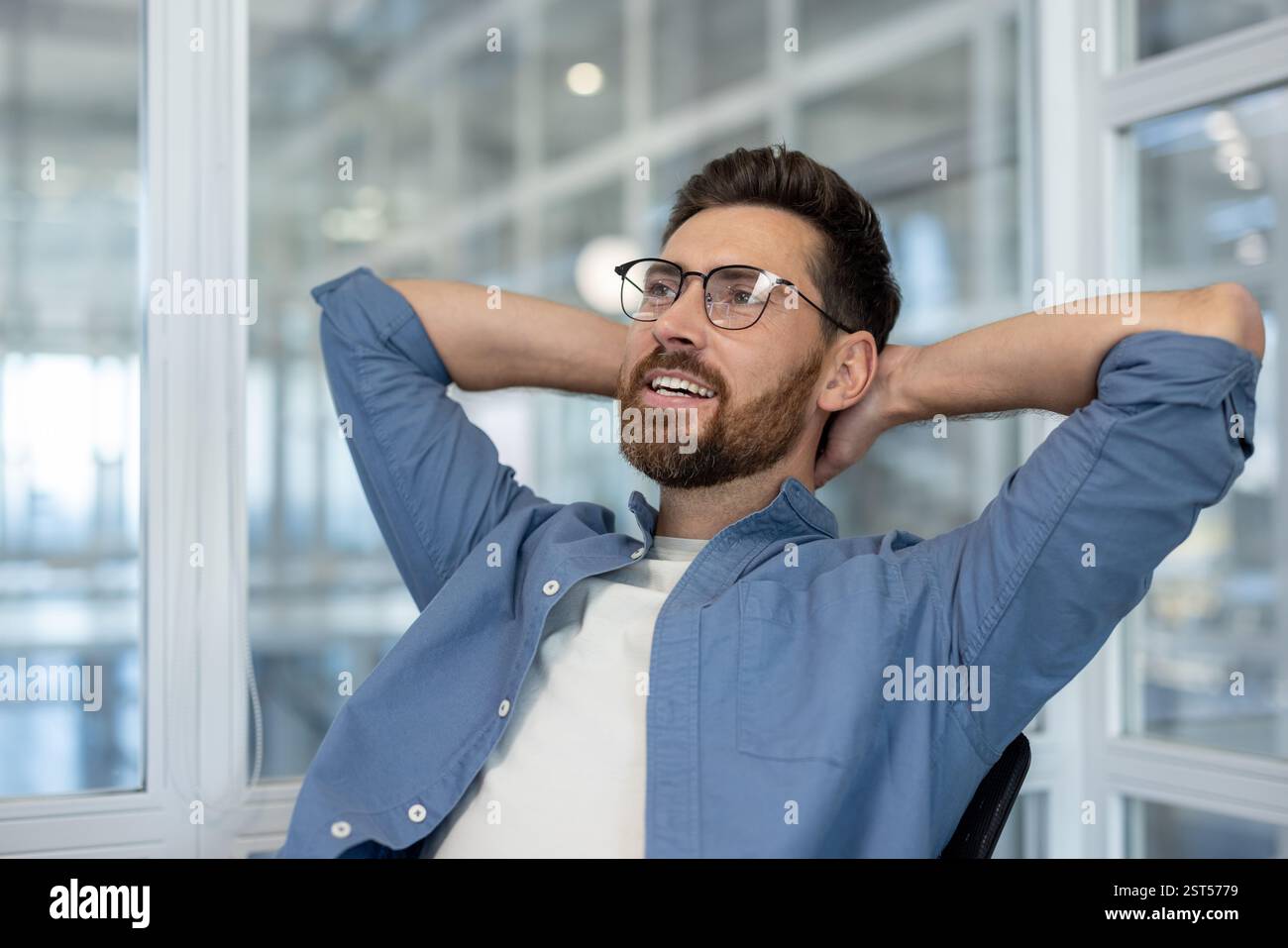 A thoughtful, bearded man relaxes with his hands behind his head, looking towards the window in a modern, bright office space Stock Photo