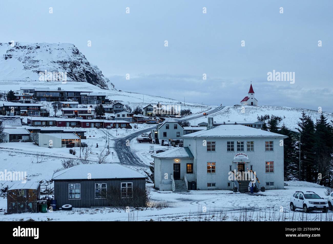 Scenic winter view of a small village covered in snow, featuring traditional houses, a winding road, and a red-roofed church on a hill. Stock Photo