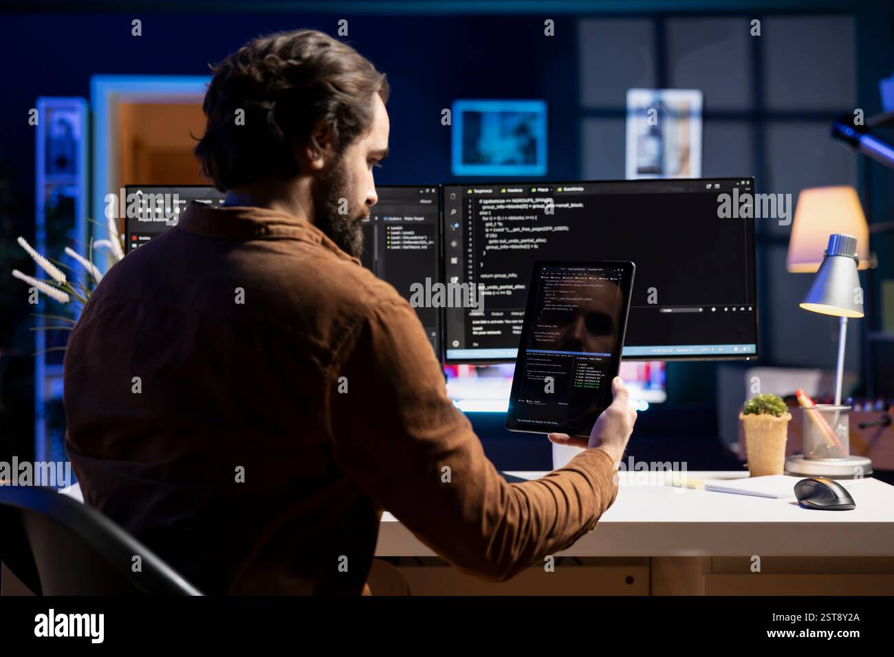 Programmer doing software debugging using tablet, checking glitches in lines of code displayed on computer monitors. Man working from home fixing coding issues using portable digital device Stock Photo