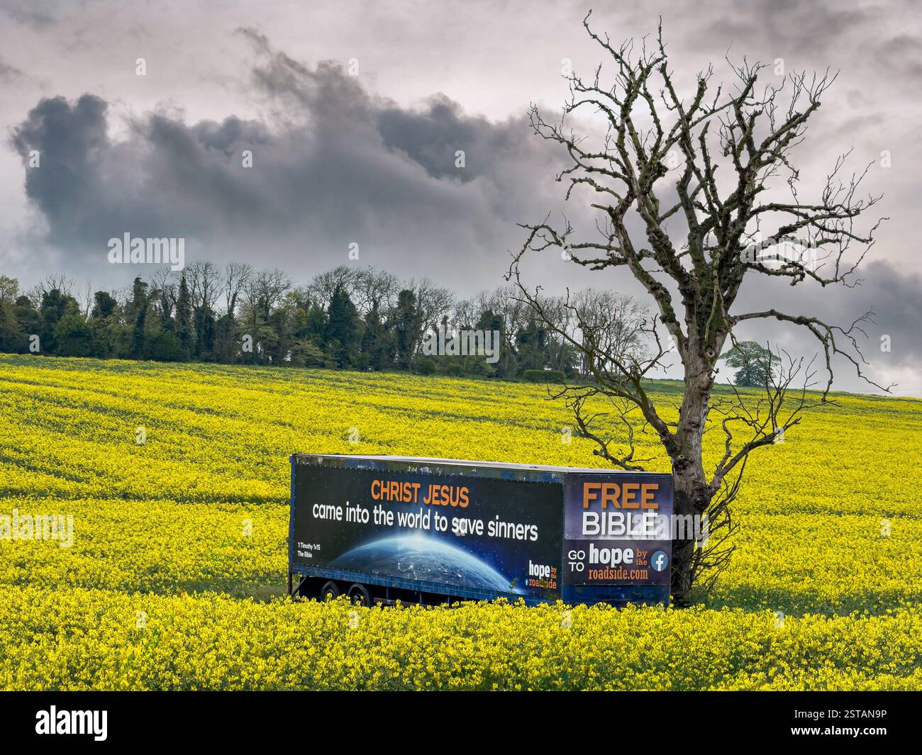 Lorry trailer with religious ad parked next to a dead tree, in a field of yellow rapeseed. Seen from the A1 in South Yorkshire, UK. Stock Photo