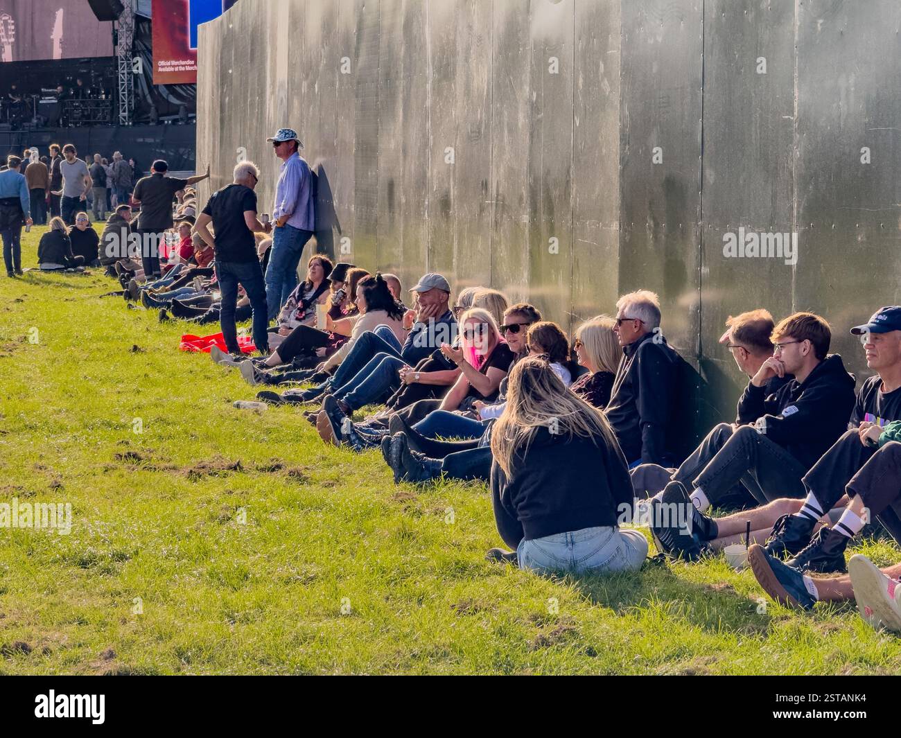 Line of people sitting on the grass, relaxing against a temporary wall in the Wythenshawe Park sunshine, waiting for a band to play Stock Photo