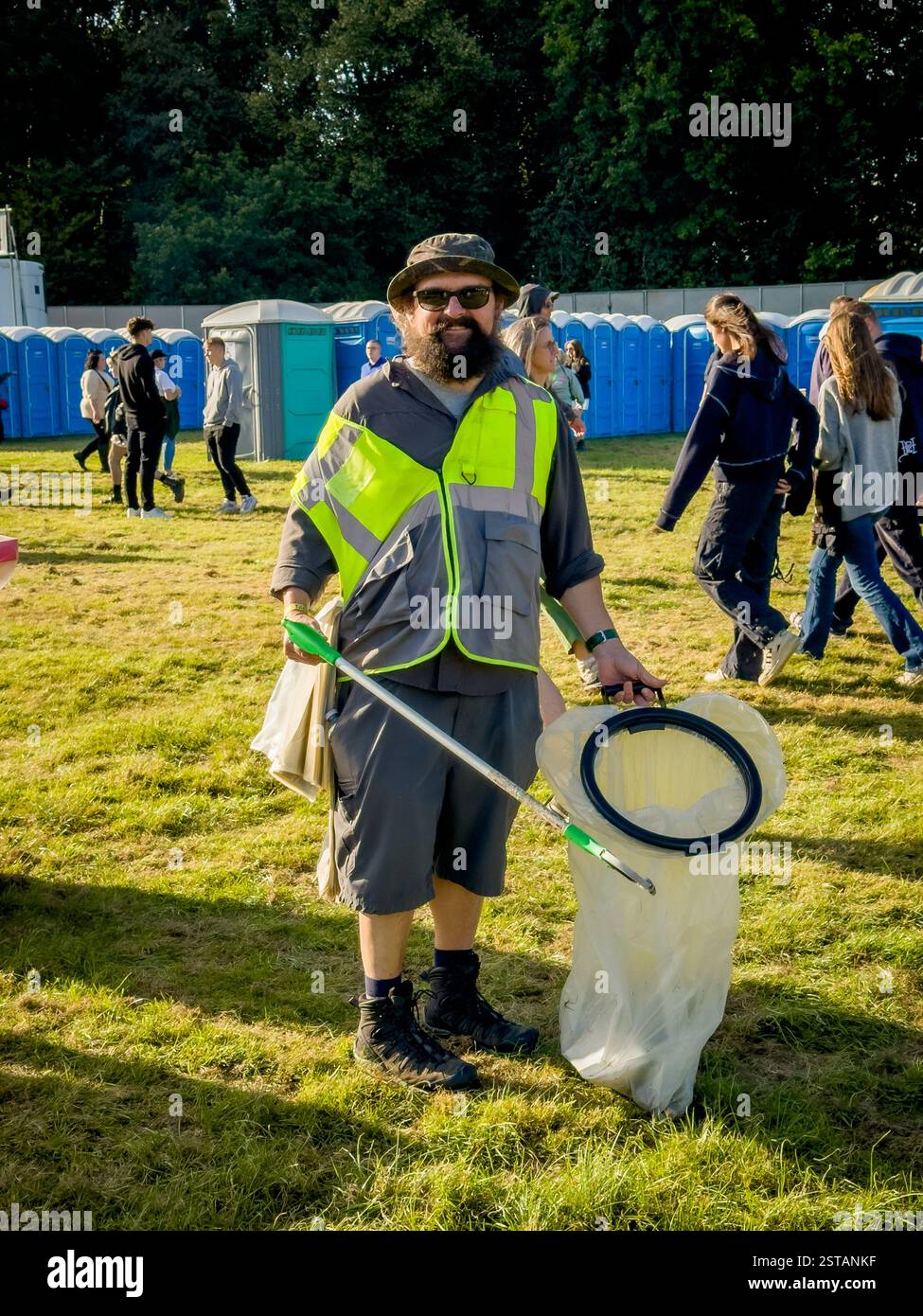 Smiling male event steward collecting litter at Wythenshawe Festival, standing in front of rows of portable toilets on a sunny evening. Stock Photo
