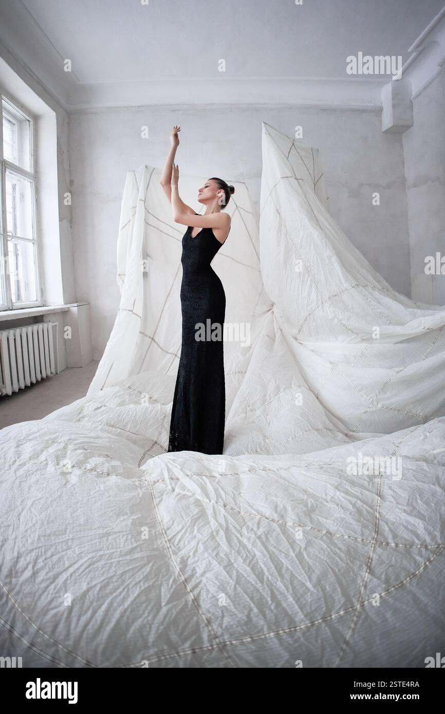 model stands gracefully in a black gown, reaching towards the ceiling. Surrounding her is an artfully arranged backdrop of white fabric, enhancing the Stock Photo