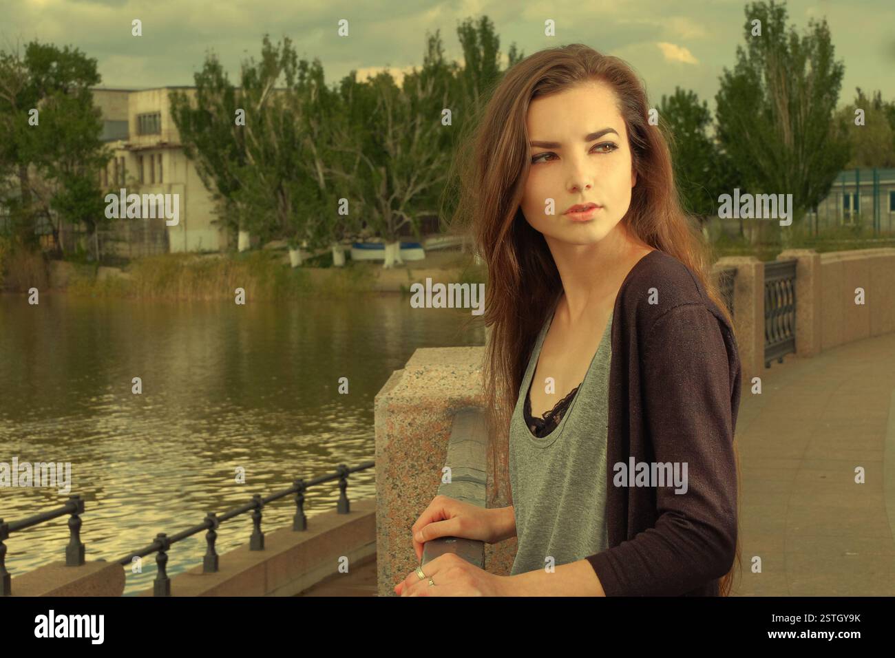 Pretty girl wearing black shirt, standing against fence by Volga River in Astrakhan in summer, looking back, filtered effect. Stock Photo